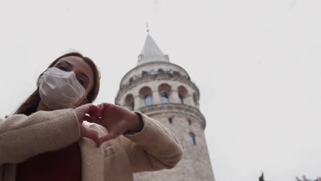 Beautiful-girl-wearing-protective-medical-mask-and-fashionable-clothes-does-heart-shape-with-hands-at-Galata-Tower-background