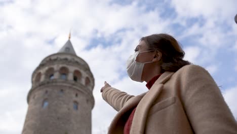Slow-Motion:Beautiful-girl-wearing-protective-medical-mask-and-fashionable-clothes-shows-sign-of-Galata-Tower-New-normal-lifestyle,-travel-concept