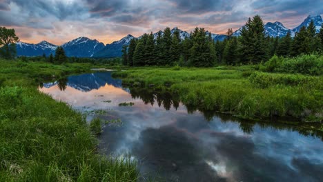 Beautiful-pink-and-orange-sunset-time-lapse-at-mirror-reflective-lake-with-snow-capped-mountains-and-thick-fluffy-clouds-flowing-in-Grand-Teton-National-Park,-Wyoming,-USA