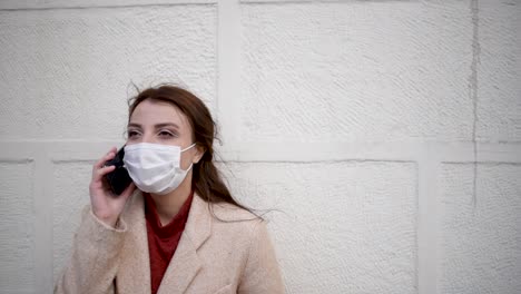 Beautiful-girl-wearing-protective-medical-mask-and-fashionable-clothes-speaks-with-a-smart-phone-in-front-of-wall