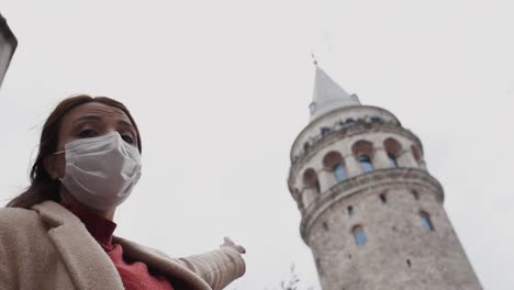 Slow-Motion:Beautiful-girl-wearing-protective-medical-mask-and-fashionable-clothes-shows-sign-of-Galata-Tower-New-normal-lifestyle,-travel-concept