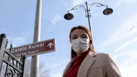 Beautiful-girl-wearing-protective-medical-mask-and-fashionable-clothes-does-heart-shape-with-hands-at-Galata-Tower-sign-background