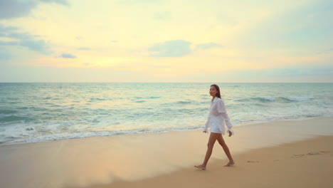 Young-girl-slowly-walking-barefoot-along-the-shore-near-the-ocean-with-waves-reaching-the-wet-sand-during-sunset