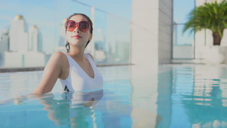 Close-up-of-a-pretty-young-woman-with-an-urban-skyline-in-the-background-relaxes-the-water-of-a-rooftop-swimming-pool