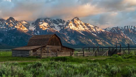 Beautiful-golden-sunrise-timelapse-at-a-farm-with-barn-and-snow-capped-mountain,-clouds-flowing-by-at-the-T