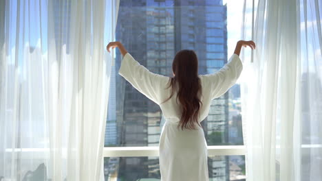 Young-Woman-in-Bathrobe-Opening-Window-Curtains-of-Bedroom-on-Sunny-Morning-With-Beautiful-View-of-City-and-Stretching-Hands