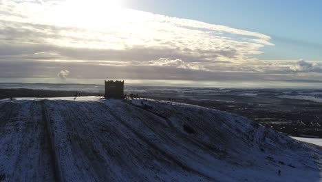 Snowy-Rivington-Pike-tower-Winter-hill-aerial-view-people-sledding-downhill-at-sunrise-slow-push-in-over-tower