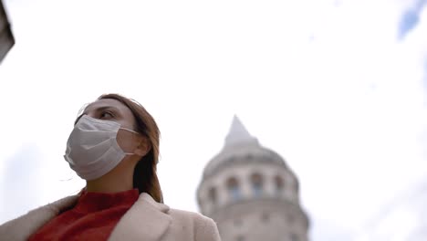 Beautiful-girl-in-fashionable-clothes-holds-and-wears-disposable-mask-poses-with-Galata-Tower-at-background