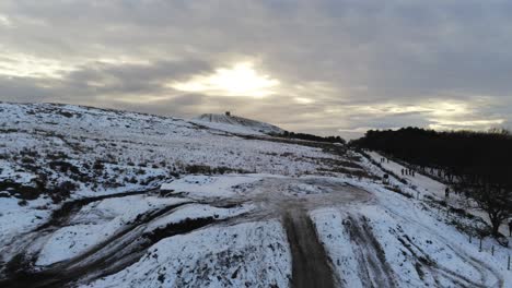 Snowy-Rivington-Pike-tower-Winter-hill-aerial-rising-view-people-sledding-downhill-at-sunrise