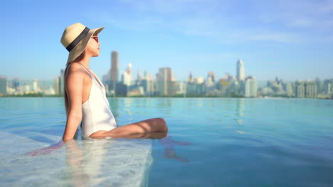 A-young-woman-sitting-in-the-water-in-an-infinity-edge-rooftop-resort-pool-looks-out-at-the-city-skyline-of-highrises