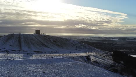Snowy-Rivington-Pike-tower-Winter-hill-aerial-view-people-sledding-downhill-at-sunrise-rising-left