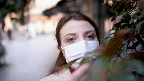 Beautiful-girl-wearing-protective-medical-mask-and-fashionable-clothes-looks-at-camera-behind-of-plants