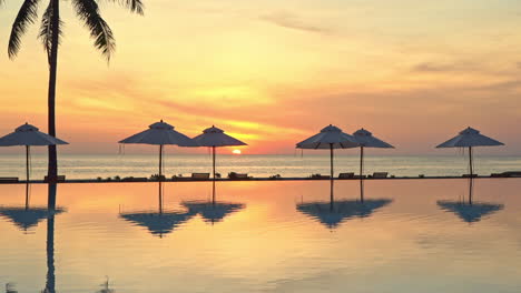 Orange-Tropical-Sunset-Above-Sea-and-Pool-of-Luxury-Resor-With-Tree-and-Parasols-Silhouettes-and-Mirror-Reflection-on-Water,-Static