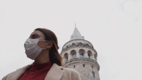 Slow-Motion:Beautiful-girl-in-fashionable-clothes-holds-and-wears-disposable-mask-poses-with-Galata-Tower-at-background