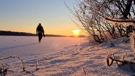 Backpacker-or-photographer-man-walking-towards-golden-sun-in-winter-wonderland,-static-low-angle-shot
