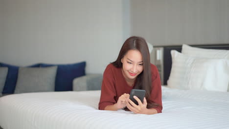 Junge-Asiatische-Frau,-Die-Auf-Dem-Bett-Liegt,-Chattet-Auf-Dem-Smartphone-Und-Lächelt