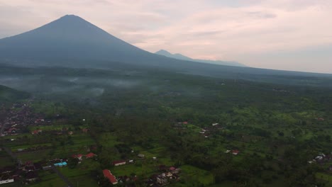 Vista-Aérea-De-Drones-Amed-Bali-Indonesia-Pequeño-Pueblo-De-Pescadores-Con-Volcán-Activo-Monte-Agung-Y-Brumoso-Vegetación-De-Selva-Brumosa-Palmera-Verde-Durante-El-Amanecer-De-La-Mañana