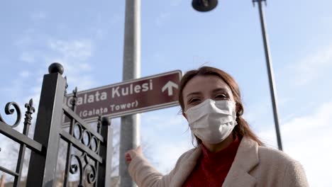 Beautiful-girl-wearing-protective-medical-mask-and-fashionable-clothes-shows-sign-of-Galata-Tower-New-normal-lifestyle,-travel-concept
