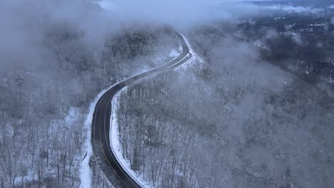 Flying-through-clouds-over-snow-covered-mountains-and-forests-in-a-mountain-valley-with-a-scenic-highway-below,-on-a-cloud-winter’s-day