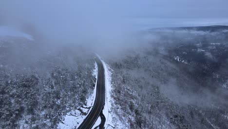 Flying-through-clouds-over-snow-covered-mountains-and-forests-in-a-mountain-valley-with-a-scenic-highway-below,-on-a-cloud-winter’s-day