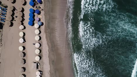 Birds-Eye-Drone-Aerial-View-of-Ocean-Waves-Breaking-on-Sandy-Beach-With-Parasols-on-Sunny-Day