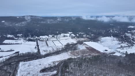 Flying-over-snow-covered-farmland-above-a-mountain-valley-on-a-cloudy-winter's-day