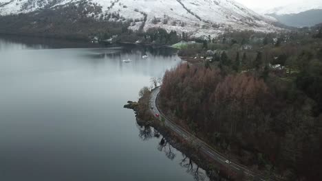 Mehrere-Autos-Fahren-Auf-Der-Linken-Straßenseite-In-Schottland-Mit-Einem-Atemberaubenden-Blick-Auf-Loch-Lomond-Mit-Der-Spitze-Des-Berges,-Der-Von-Weißem-Schnee-Bedeckt-Ist