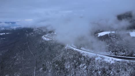 Fliegen-Durch-Wolken-über-Schneebedeckte-Berge-Und-Wälder-In-Einem-Bergtal-Mit-Einer-Malerischen-Autobahn-Darunter,-An-Einem-Wolkenwintertag