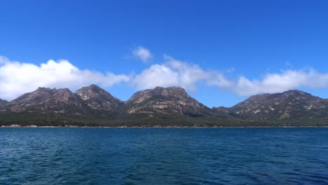 Timelapse-of-white-clouds-rolling-over-rocky-mountain-range-on-sunny-clear-summer-day-with-blue-ocean-water-in-foreground,-Freycinet,-Tasmania