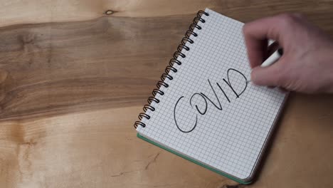 Man-Writing-COVID-19-On-Graph-Paper-Using-A-Marker-On-Top-Of-Wooden-Table