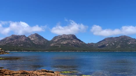 Timelapse-of-clouds-rolling-over-rocky-mountain-range-on-sunny-clear-summer-day-with-blue-ocean-water-trickle-in-foreground,-Freycinet,-Tasmania