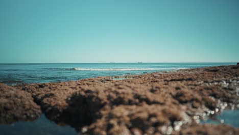 Ocean-shoreline-flat,-with-cargo-boats-and-waves-hit-rocks-softly-at-sunshine-wide-angle-4K