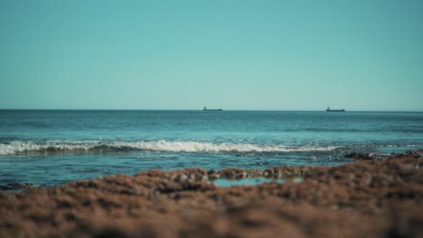 Ocean-shoreline-flat,-with-cargo-boats-away-and-waves-hit-rocks-softly-at-sunshine-4K