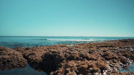 Ocean-shoreline-with-flat-cargo-boats-away,-waves-hit-rocks-gently-out-of-focus-at-sunshine-4K
