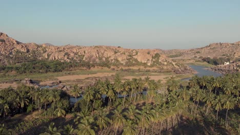 Paradisiac-Tungabhadra-Riverbank-among-rocky-landscape-and-lush-palm-trees-in-Hampi,-Karnataka,-India---Aerial-panoramic-wide-shot