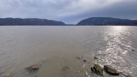 Side-pan-river-beach-hyper-lapse-during-winter,-with-distant-mountains-lightly-covered-in-snow-and-a-rocky-shoreline