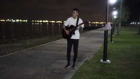 Hombre-Tocando-La-Guitarra-Solo-Por-La-Noche-En-El-Parque-De-La-Costa-Este,-Singapur-Mientras-Está-Oscuro-Con-La-Luz-De-Los-Barcos-Y-La-Luz-De-La-Calle