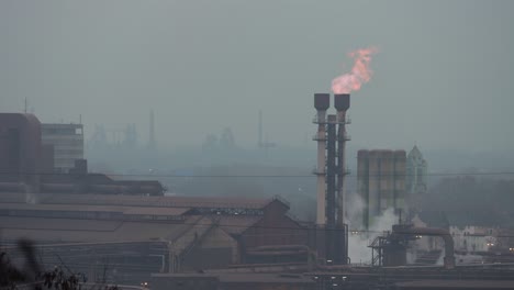 Red-and-blue-flames-burning-out-of-a-chimney-of-a-factory-with-more-plants-on-the-background-on-a-cloudy-grey-day-in-the-ruhr-area