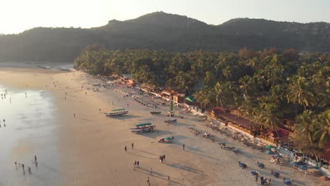 Low-tide-Palolem-Beach-at-sunset-with-tourists-enjoying-the-paradisiac-ocean-shore,-in-Goa,-India--Aerial-Low-angle-Slow-Orbit-shot