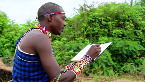 Hombre-Masai-Escribiendo-Notas-En-Un-Libro,-Sentado-Al-Aire-Libre,-En-Kenia,-África