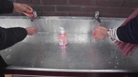Two-girls-washing-hands-with-soap-and-water-in-metallic-sink-outside