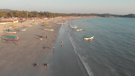 Tourists-strolling-along-the-golden-sands-near-the-coastline-with-moored-fishing-boats-in-Palolem-Beach,-in-Goa,-India---Aerial-Fly-over