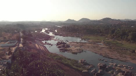 Tungabhadra-River-flow-cascading-alongside-the-rocky-landscape-Hampi-town's-edge-in-Karnataka,-India---Aerial-Fly-over