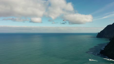 Distant-paraglider-flies-over-open-ocean-close-to-shore-of-Madeira,-aerial