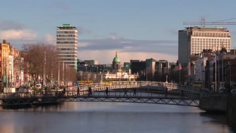 Still-shot-wide-of-people-walking-over-a-bridge-with-iconic-Dublin-building-and-some-transport-vehicles-passing-by-day-time