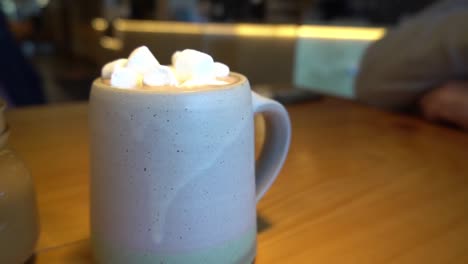 1-3-Bokeh-backgroud-at-coffee-shop-with-employees-making-drinks-for-customers-ordering-beverages-and-in-the-forground-a-jar-shaped-late-hot-chocolate-with-blurred-people-seated-at-rotating-table-view
