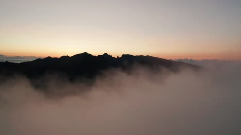 Rising-out-of-clouds-revealing-silhouette-mountain-during-dawn,-aerial
