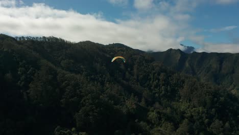 Paraglider-gliding-in-air-above-lush-forest-mountains-of-Madeira-island,-aerial