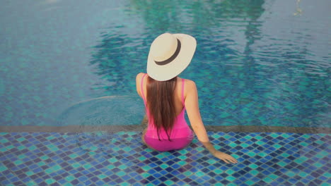 Back-view-of-young-woman-with-large-hat-sitting-in-pool-water