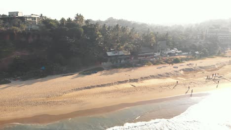Tropical-golden-sandy-beach-bathed-by-the-warm-waters-of-the-Arabian-sea-in-Varkala,-Kerala,-India---Aerial-low-angle-panoramic-shot